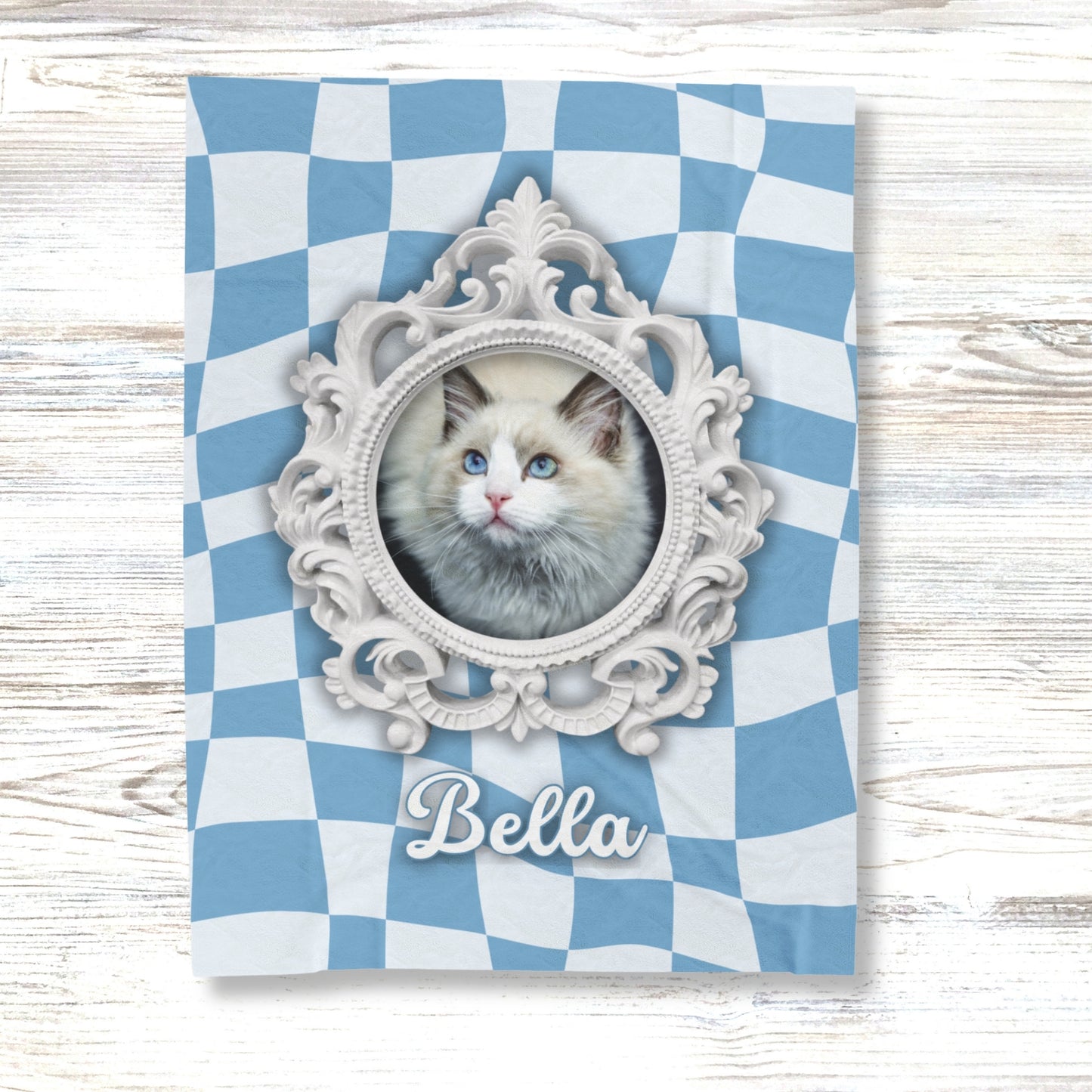Custom cat blanket with ornate white frame, blue checkered background, and name "Bella" displayed on light wood surface