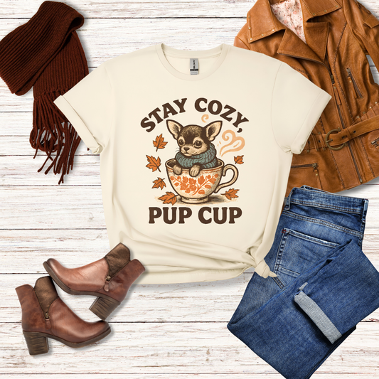 Flat lay of natural color “Stay Cozy Pup Cup” Chihuahua graphic t-shirt styled with jeans, boots, scarf, and jacket for fall fashion