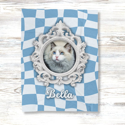 Custom cat blanket with ornate white frame, blue checkered background, and name "Bella" displayed on light wood surface